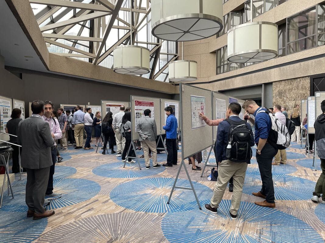 People view and discuss research posters in a large, well-lit atrium with geometric flooring. Some participants take notes, while others engage in conversations in front of the display boards.