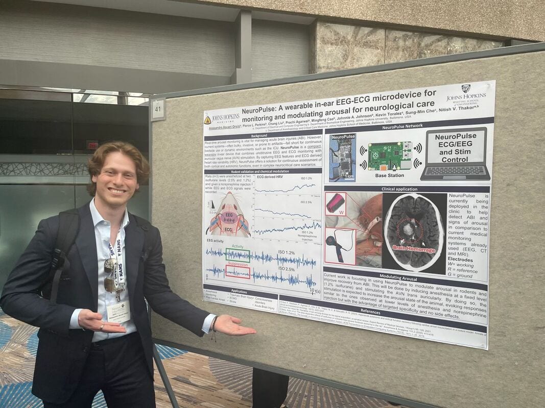 A man in a suit smiles and gestures toward a scientific poster about a wearable in-ear EEG-ECG microdevice for neurological care, displayed on a bulletin board at a conference.