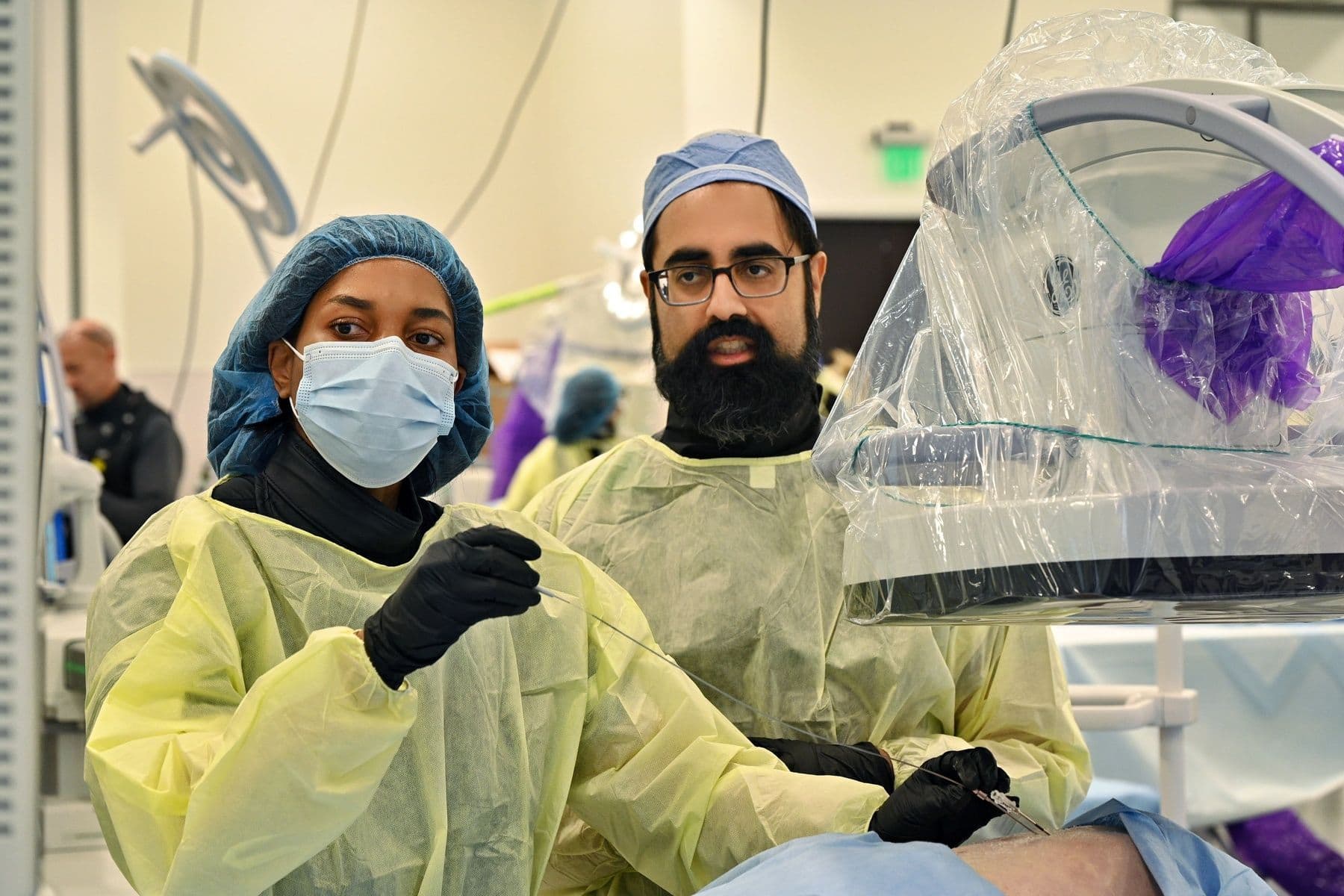 Two medical professionals in scrubs and masks perform a procedure in a hospital room, using medical equipment. One holds a syringe while the other observes.