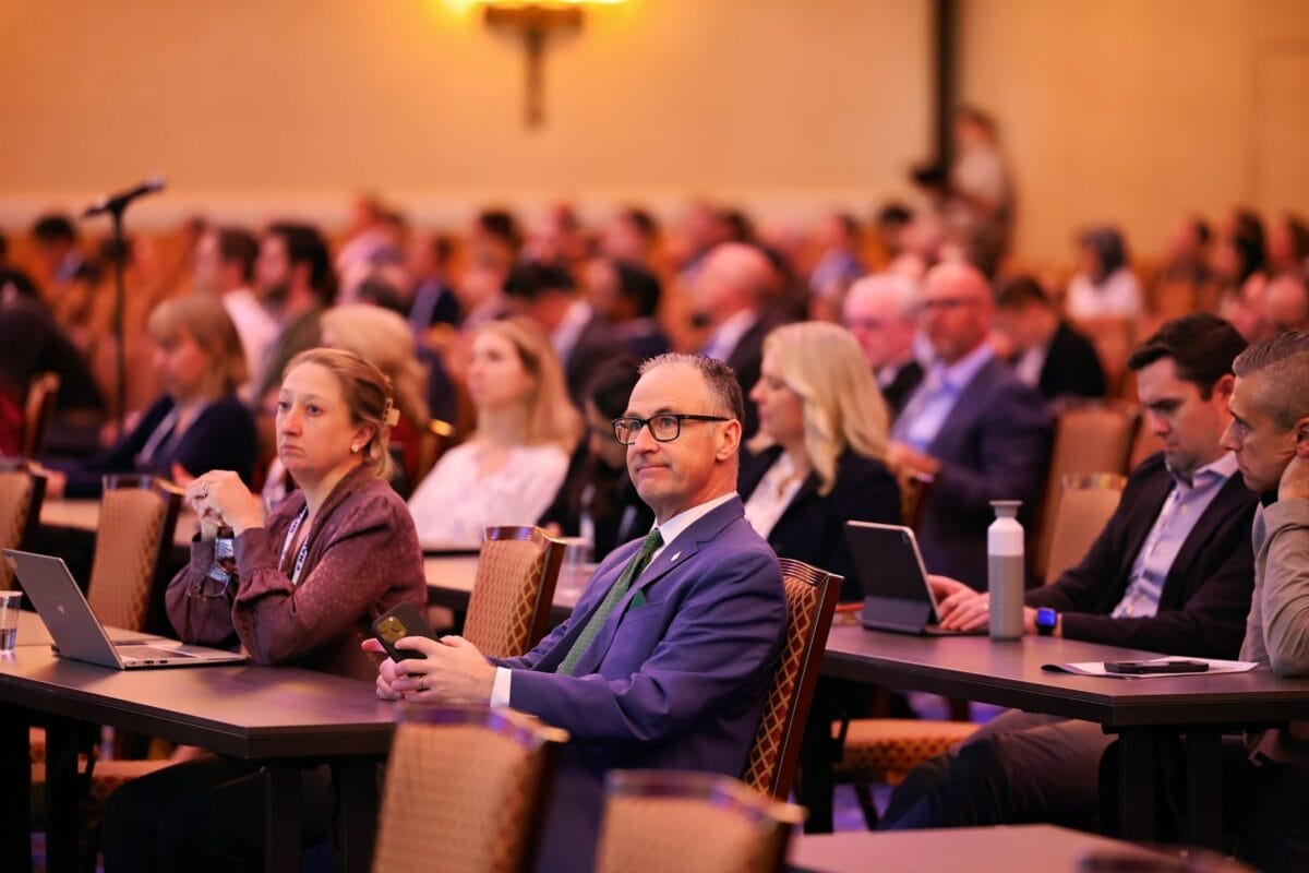 A large group of people in business attire sit at tables during a conference, listening attentively, with laptops and notebooks open in front of them.