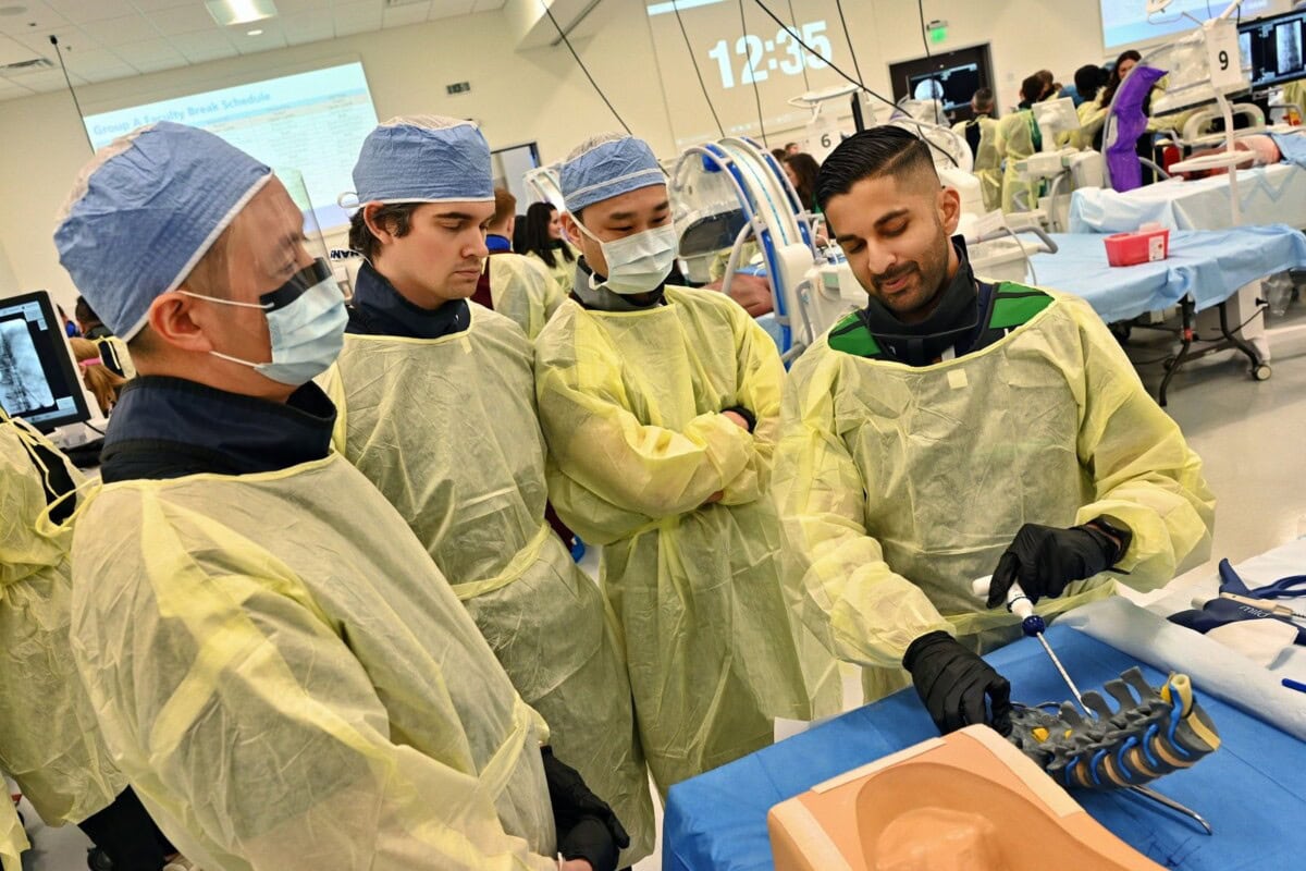 Four people in medical gowns and masks observe a medical procedure demonstration on a mannequin arm in a clinical training room with medical equipment and monitors in the background.