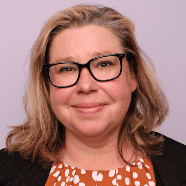 A person with shoulder-length blonde hair, wearing glasses, a black blazer, and an orange shirt with white spots, smiles softly in front of a plain light purple background.