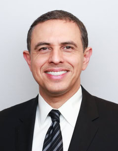 Smiling man in a dark suit, white shirt, and striped tie with short dark hair, posing against a plain light background.
