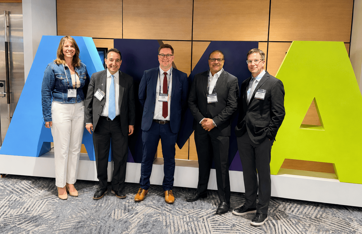 Five people in business attire stand smiling in front of large, colorful AMA letters on a modern office floor with light wood and blue decor.
