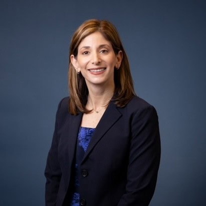A woman with straight brown hair, wearing a dark blazer over a blue top, smiles while posing against a plain dark blue background.