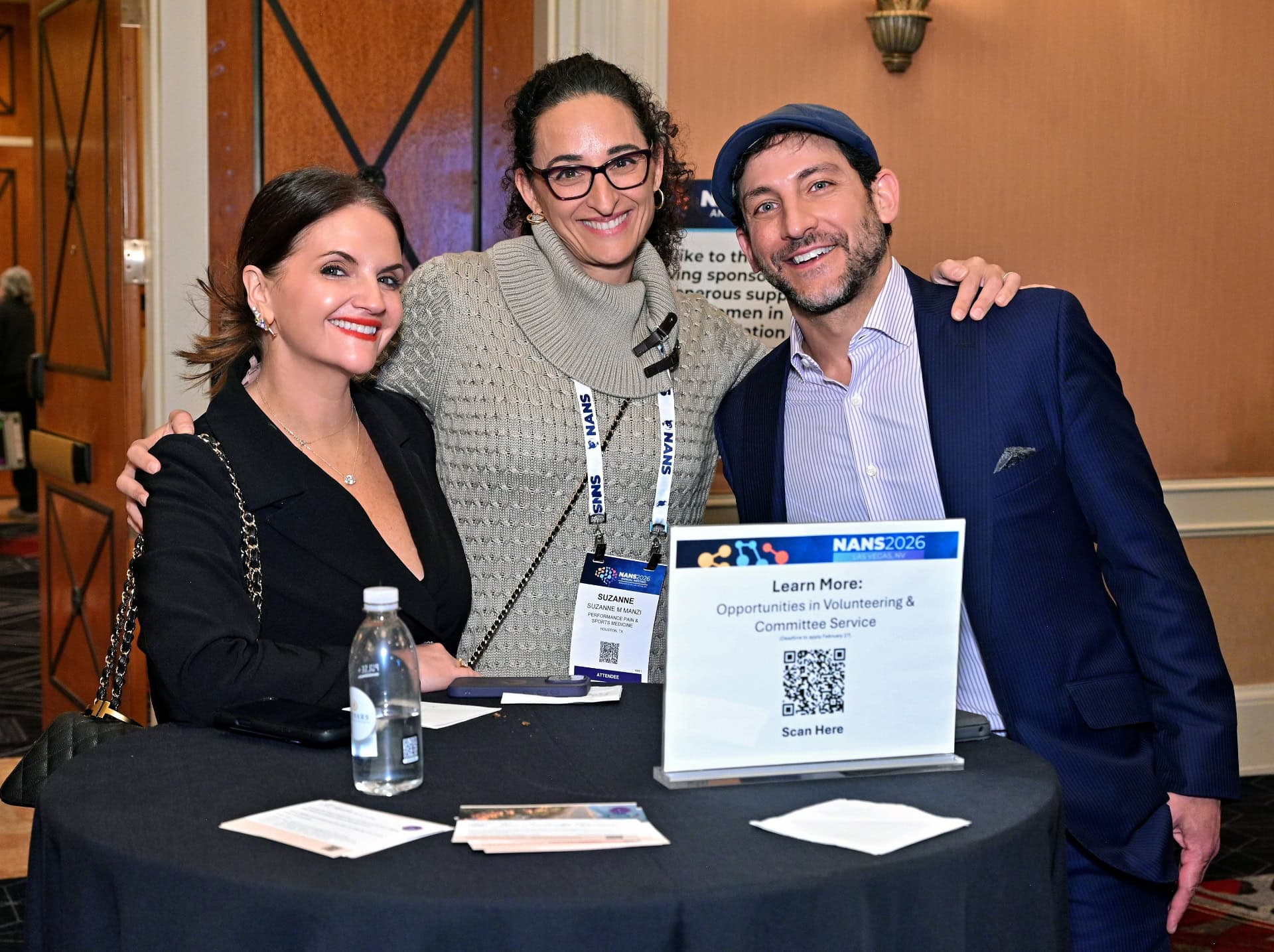 Three people smile at a round table with a water bottle and a sign reading Learn More: Opportunities in Volunteering & Committee Service. They appear to be at a professional event or conference.