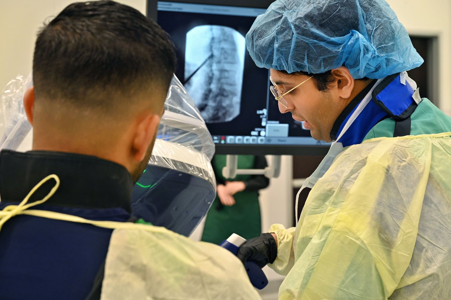 Two medical professionals in protective gear perform a procedure, looking at a spine X-ray on a monitor in a clinical setting.