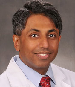 A smiling man wearing a white medical coat, blue shirt, and red plaid tie, posing in front of a neutral gray background.