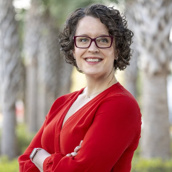 Woman with short curly brown hair and glasses, wearing a red blouse, smiling with arms crossed, standing outdoors in front of blurred palm trees.