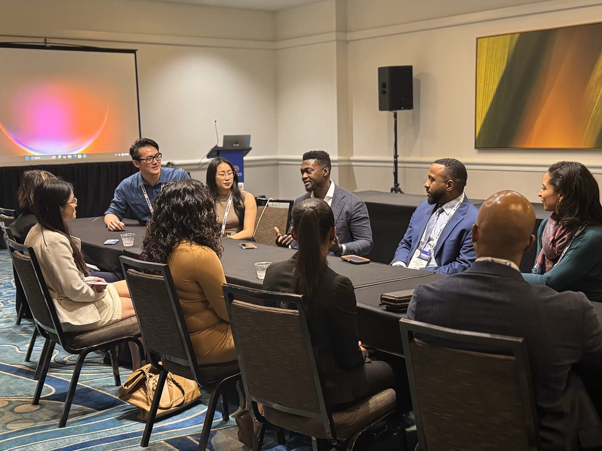 A diverse group of professionals sit around a conference table in discussion, some smiling, with a screen and abstract artwork in the background.