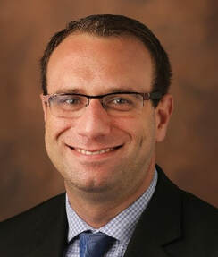 A man wearing glasses, a suit, and a blue tie smiles at the camera against a brown, blurred background.