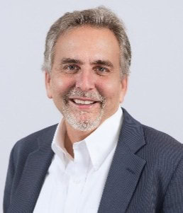 Smiling middle-aged man with short gray hair, a trimmed beard, and mustache, wearing a dark blazer over a white collared shirt, posing in front of a plain light background.
