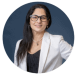 Woman with long dark hair, wearing glasses, a white blazer, black top, pearl necklace, and earrings, smiles at the camera against a plain dark blue background.