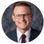 A man wearing glasses, a navy suit, white shirt, and red-striped tie smiles in a professional headshot with a gray textured background.