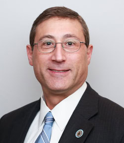 A man wearing glasses, a dark suit, white shirt, and striped tie, smiling in front of a plain light background.