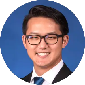 A man with short dark hair and glasses, wearing a dark suit, white shirt, and blue tie, smiling in front of a solid blue background.