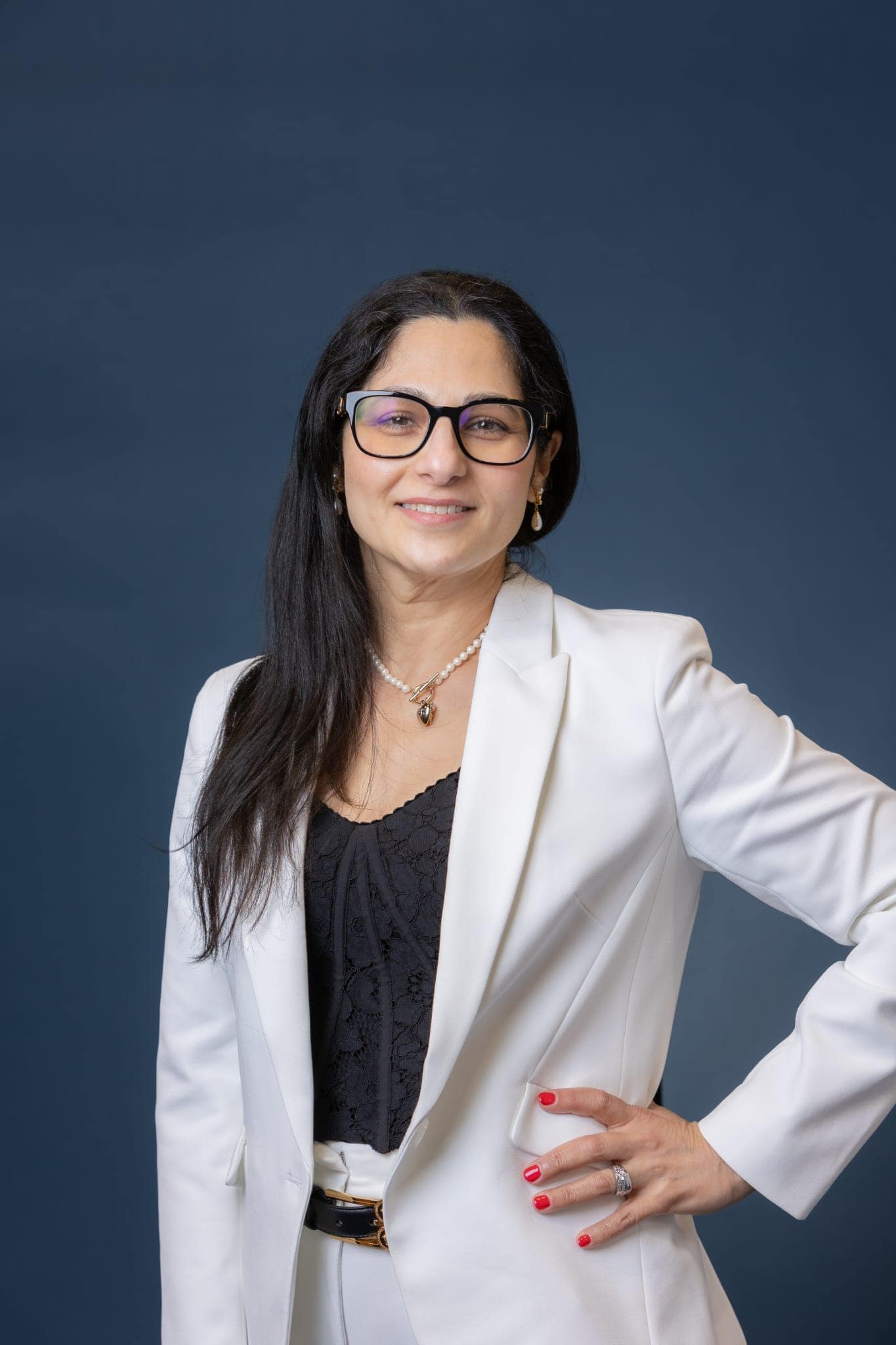 A woman with long dark hair, glasses, and a white suit stands confidently against a solid dark blue background, smiling with her hand on her hip.