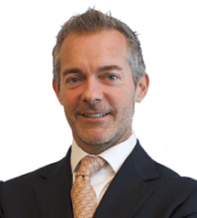 A man in a dark suit and patterned tie smiles slightly, standing against a white background. He has short, grayish hair and light facial stubble.