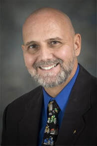 A smiling bald man with a gray beard wearing a blue shirt, a dark suit jacket, and a colorful patterned tie, posing in front of a neutral background.