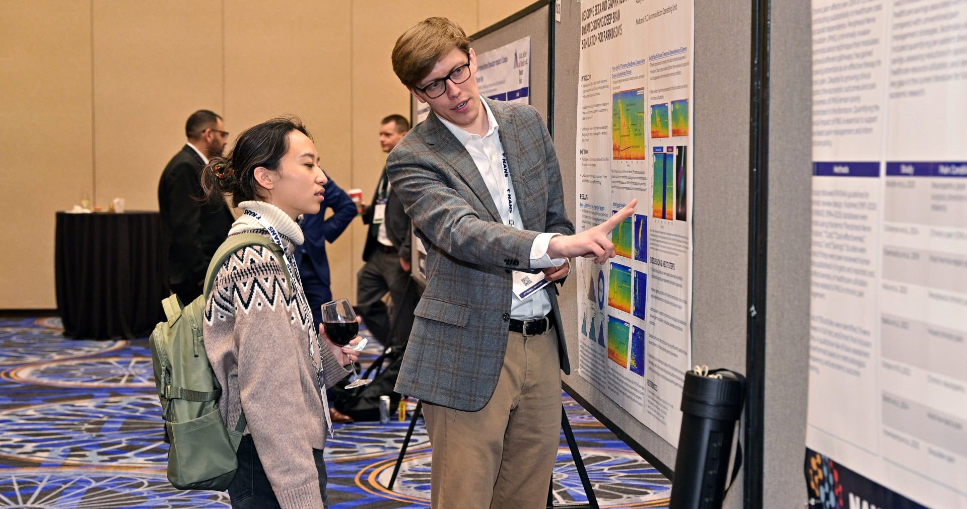 A man in a blazer explains a scientific poster to a woman holding a drink at a conference. Other attendees are seen talking in the background.
