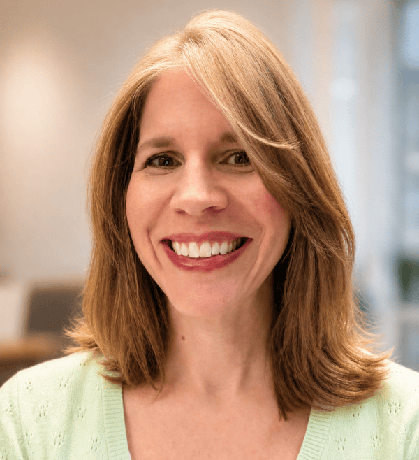 A woman with shoulder-length light brown hair smiles at the camera. She is wearing a light green sweater, and the background is softly blurred, suggesting an indoor setting.