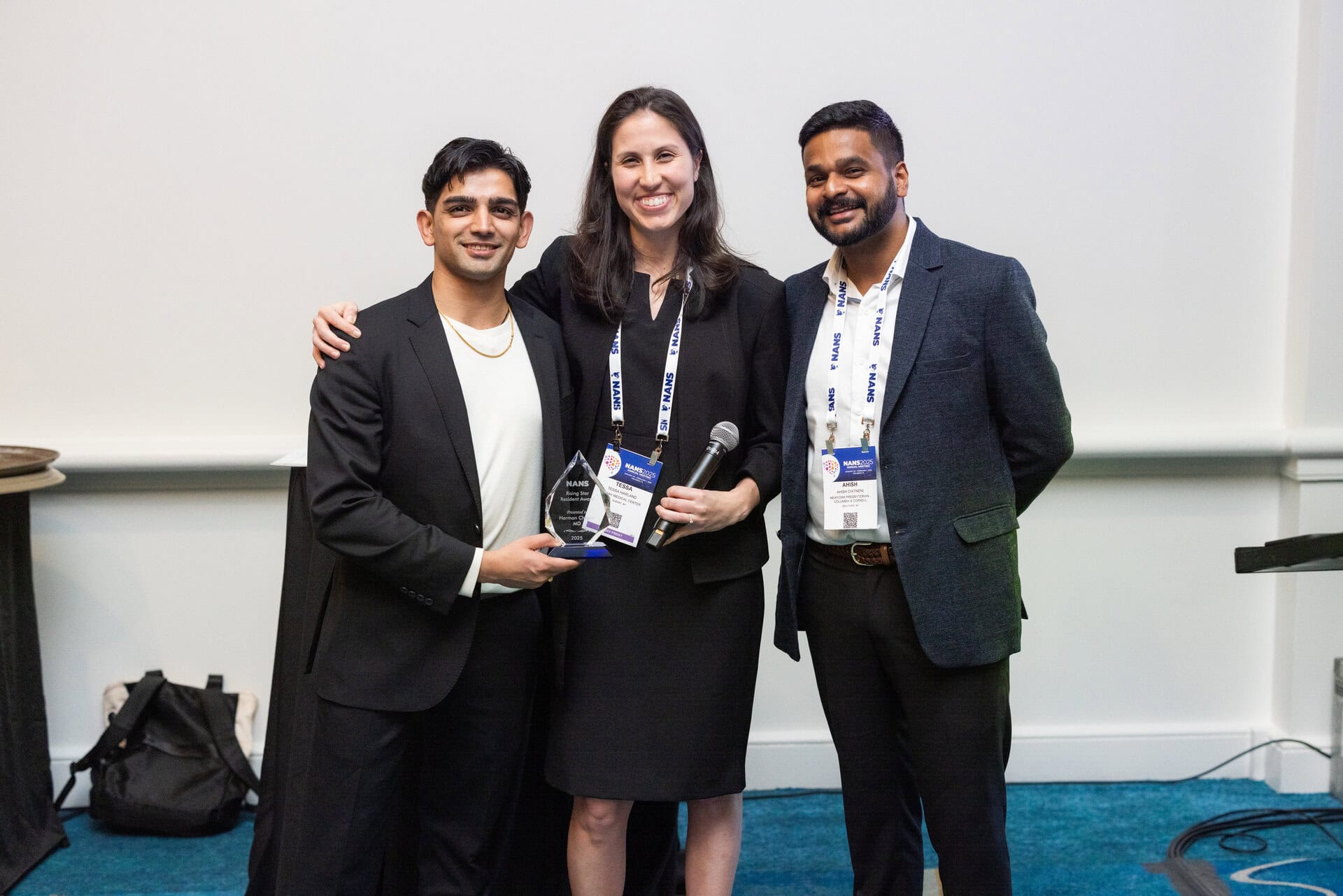 Three people in business attire stand together at an event. One holds a glass award, another holds a microphone, all are smiling. They have conference badges and are posing against a plain white wall.