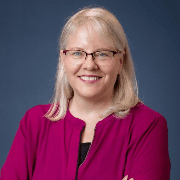 A woman with blonde hair and glasses, wearing a magenta blouse, smiles with arms crossed against a solid blue background.