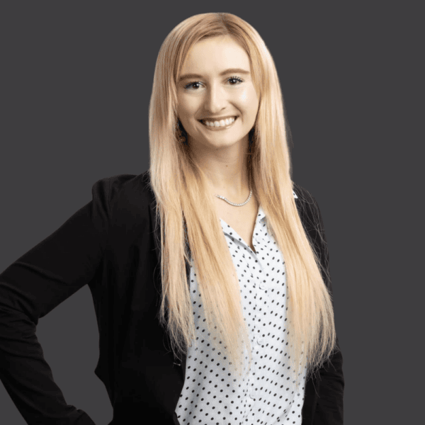 Smiling woman with long blonde hair wears a black blazer over a white blouse with black polka dots, standing against a plain dark gray background.