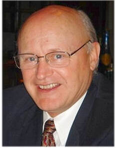 A smiling older man with glasses, a bald head, and wearing a dark suit jacket, white shirt, and patterned tie.