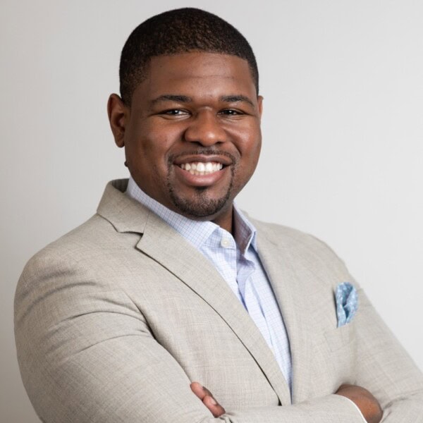 A man in a light beige suit jacket and light blue shirt smiles confidently with arms crossed in front of a plain light background.