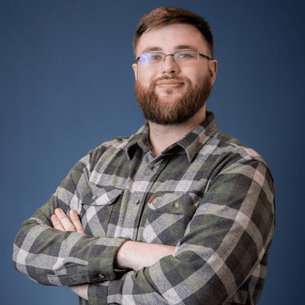 A bearded man with glasses wearing a green and gray plaid shirt stands with arms crossed, smiling confidently in front of a plain blue background.