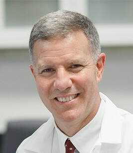 A middle-aged man with short gray hair wearing a white lab coat and a red tie smiles at the camera, set against a soft-focus indoor background.