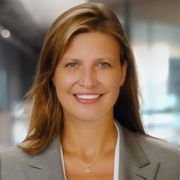 A woman with straight, light brown hair wearing a gray blazer and a delicate necklace smiles in an office setting with blurred background.
