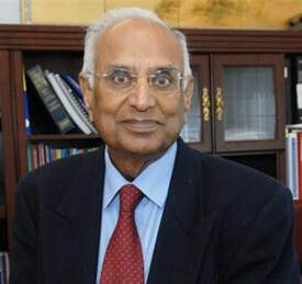 An older man with glasses, gray hair, and a suit with a red tie, sitting in an office with bookshelves in the background.
