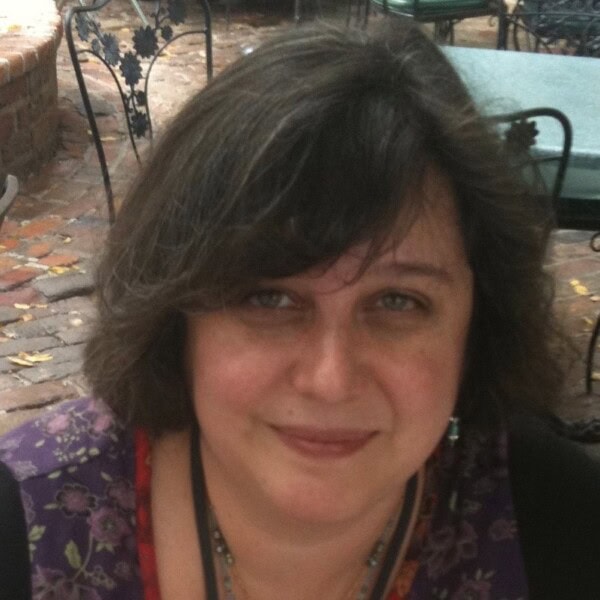 A woman with shoulder-length brown hair sits outdoors, smiling softly. She wears a floral-patterned top and a black necklace. Metal chairs and a brick patio are visible in the background.