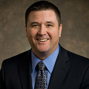 Man in a dark suit, blue shirt, and patterned tie, smiling at the camera against a brown mottled background.