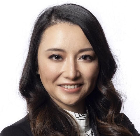 Woman with long, wavy dark hair and light skin smiles at the camera. She is wearing a black blazer over a white patterned top and poses in front of a plain white background.