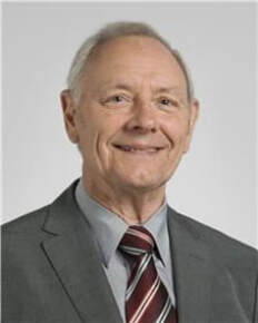 Smiling older man with short gray hair wearing a gray suit, striped tie, and light gray shirt, posing in front of a plain light background.
