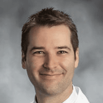 A man with short brown hair and light skin smiles at the camera, wearing a white collared shirt against a neutral, blurred gray background.