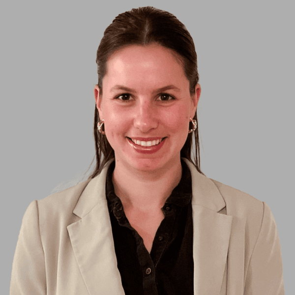 Woman with long brown hair, wearing a light blazer over a dark shirt, smiles at the camera against a plain gray background.