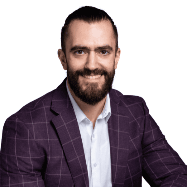 A man with a beard and slicked-back hair, wearing a purple plaid suit jacket and white shirt, smiles at the camera against a plain white background.