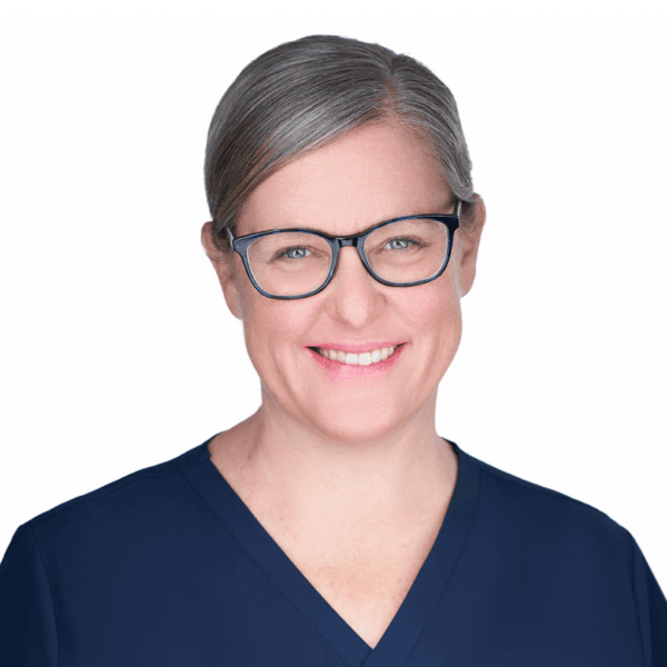Smiling woman with gray hair, glasses, and blue scrubs poses against a plain white background.