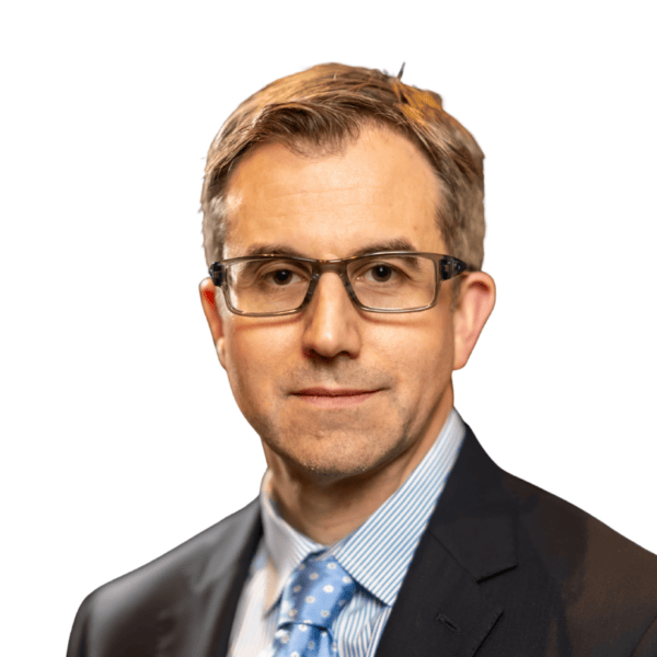 A man wearing glasses, a dark suit, a blue dress shirt, and a blue polka dot tie, posing in front of a white background.