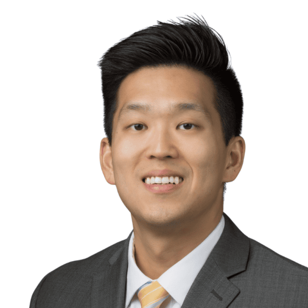 A man with short black hair wearing a gray suit, white shirt, and light yellow striped tie smiles at the camera against a plain white background.