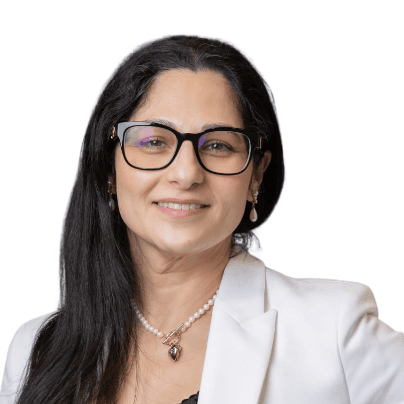 A woman with long dark hair, glasses, and a white blazer smiles at the camera. She is wearing a pearl necklace and earrings. The background is plain white.