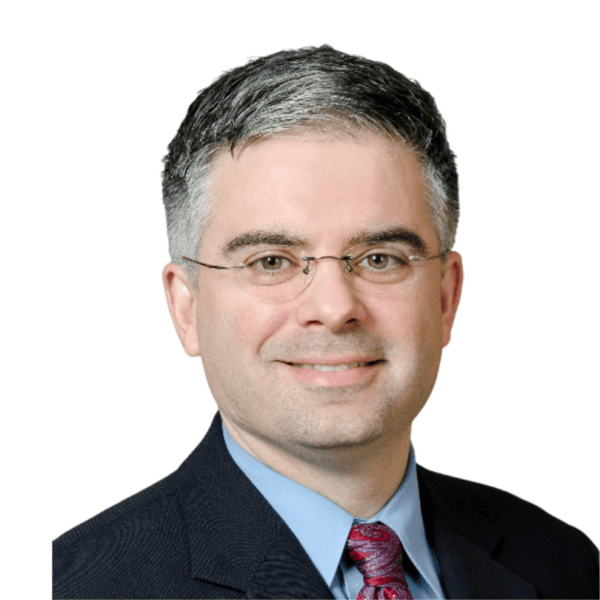 A man with short gray and black hair, wearing glasses, a dark suit, blue shirt, and red patterned tie, smiling at the camera with a white background.
