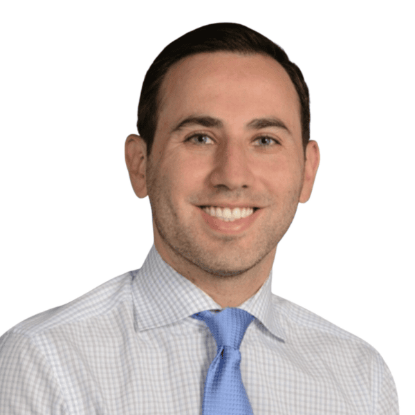 A man with short dark hair, wearing a light-colored checkered shirt and a blue tie, smiles at the camera against a plain white background.