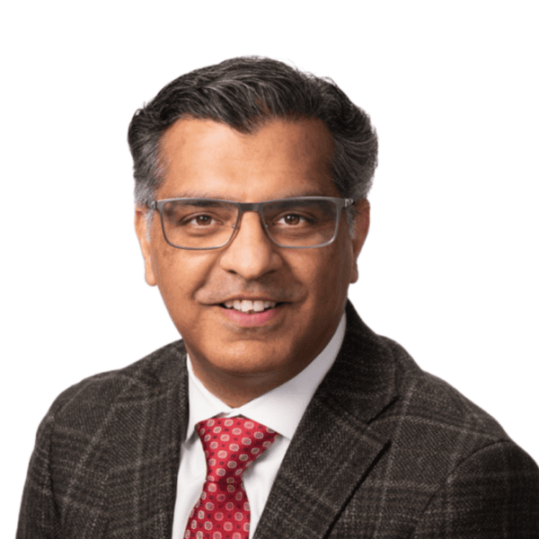 Smiling man with dark hair and glasses wearing a checkered suit jacket, white shirt, and red patterned tie on a white background.