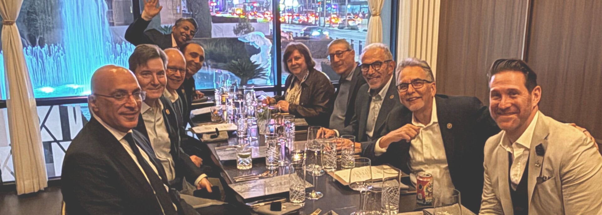 A group of people in formal attire are seated around a restaurant table, smiling at the camera. A lit fountain and city lights are visible through large windows behind them.