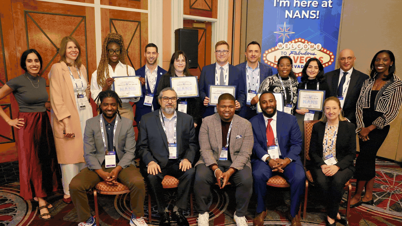 A diverse group of 16 people, some holding certificates, pose in a conference room with a blue Im here at NANS! banner in the background. Most are standing, while five are seated in front, all smiling.